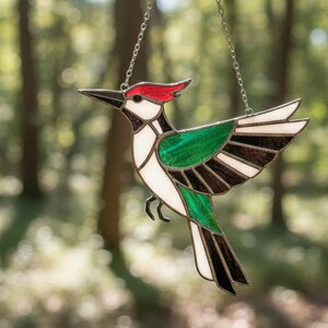 Può includere: Un acchiappasole in vetro colorato a forma di picchio con cresta rossa, faccia bianca e ali verdi e nere. L'uccello è costruito con singoli pezzi di vetro uniti da linee di saldatura in metallo scuro e pende da una sottile catena di metallo argentato.