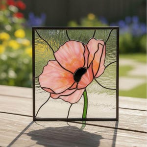 Puede incluir: Un panel de vidrieras con una flor de amapola rosa con centro negro y tallo verde. La flor se encuentra sobre un fondo transparente con líneas negras, enmarcada en un borde marrón oscuro. El panel se muestra sobre una superficie de madera.