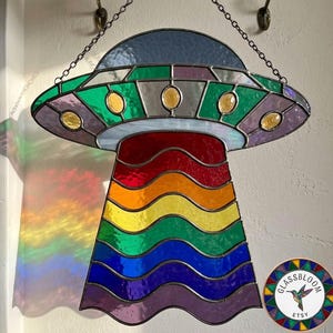 May include: Stained glass UFO suncatcher with a rainbow beam. The spacecraft has a blue top, green and purple trim, and yellow accents. The rainbow beam is made of red, orange, yellow, green, blue, and purple glass. It hangs from a chain.