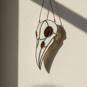 Op de afbeelding: Gebrandschilderde raven schedel wanddecoratie. De witte glazen schedel heeft rode accenten in de oogkassen en een cirkelvormig ontwerp. Opgehangen aan een zwart koord tegen een witte muur, met schaduwen.