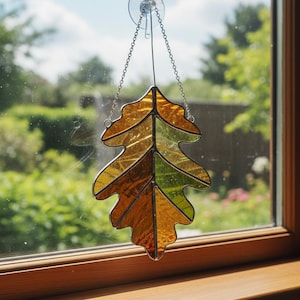 May include: A stained glass suncatcher shaped like an oak leaf, featuring segments of amber, orange, and green glass held together by dark metal solder lines. The leaf hangs from a silver-toned metal chain attached to a clear suction cup.
