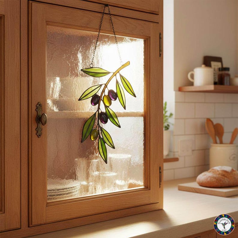 May include: A stained glass olive branch with green leaves and purple and yellow olives hangs in front of a cabinet window. The decorative piece is suspended by a delicate chain, adding a touch of nature-inspired art to the kitchen.