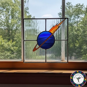 May include: Stained glass window panel featuring a blue planet with a ring and a comet in shades of orange and yellow. The panel is square, with a black border and is hanging in front of a window. The window is made of textured glass.