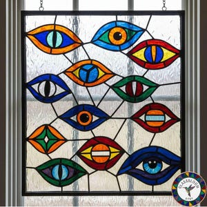 May include: Stained glass window panel featuring a geometric design of colorful eyes. The eyes are in various colors, including blue, orange, green, and red, with black outlines. The panel is framed in black and hangs from chains.
