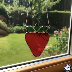 May include: A stained glass strawberry with a red body and green leaves, hanging from a silver chain. The strawberry is displayed in front of a window with a blurred outdoor background.