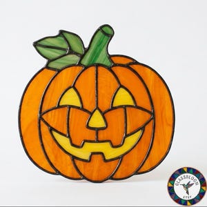 May include: A stained glass jack-o'-lantern pumpkin ornament featuring orange glass segments, yellow glass for the eyes, nose, and mouth, and a green glass leaf and stem, all held together by dark metal solder lines in a decorative suncatcher style.