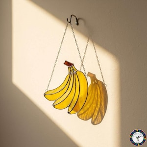 May include: A stained glass banana bunch, hanging from a chain. The yellow bananas have black outlines and a red-brown stem. The sunlight casts a shadow of the bananas on the wall. The piece is suspended from a decorative hook.