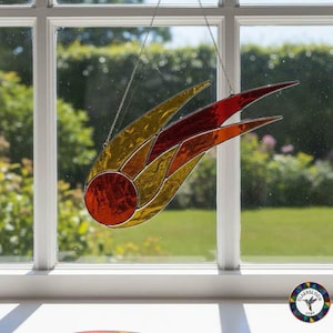 May include: A stained glass comet in shades of red, orange, and gold. The comet has a circular red centre and a trailing tail. It hangs from a chain in front of a window, creating a celestial theme. The comet's dimensions are approximately 25 cm in length.