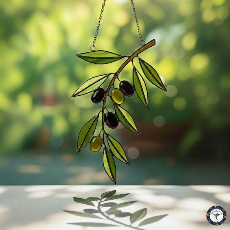 May include: Stained glass olive branch with green leaves and black and green olives, hanging from a chain. The artwork is set against a blurred green background, creating a natural and elegant aesthetic.