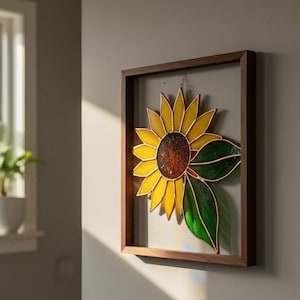 Puede incluir: Un girasol de vidriera enmarcado con pétalos amarillos, un centro marrón y hojas verdes. La obra de arte está encerrada en un marco de madera marrón oscuro y cuelga de una pared gris claro. El girasol es una pieza decorativa.