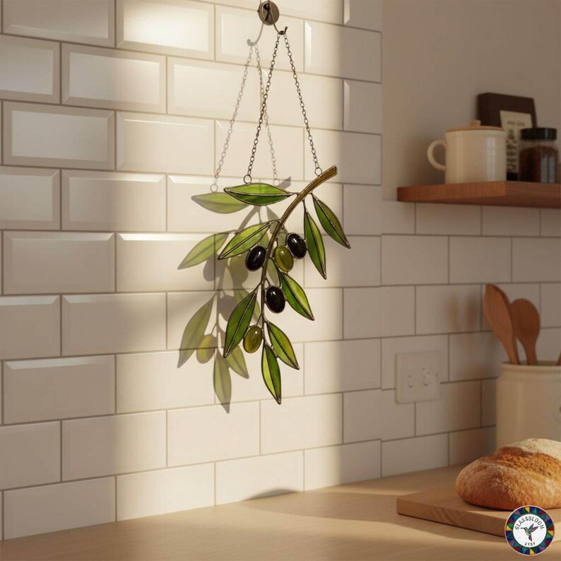 May include: A stained glass olive branch with green leaves and black and green olives hangs from a chain. The decorative piece is suspended in front of a white tiled wall, with sunlight casting shadows.