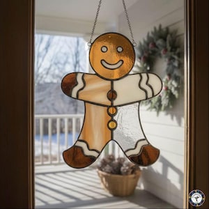 May include: A stained glass gingerbread man ornament with a smiling face, hanging from a chain. The ornament features shades of brown, white, and gold, with a decorative border. The ornament is hanging in front of a window.