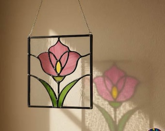 Puede incluir: Un atrapasoles cuadrado de vidrieras con un tulipán rosa, centro amarillo y hojas verdes, enmarcado en metal negro. La pieza cuelga de una fina cadena plateada y proyecta una colorida sombra floral sobre la pared que se encuentra detrás.