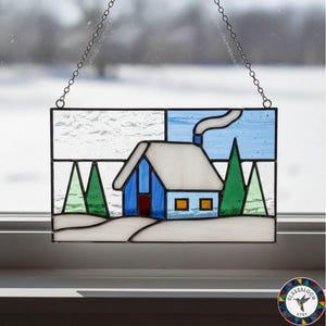 May include: Stained glass panel depicting a winter scene with a blue house, white roof, and green trees. The panel is rectangular and hangs from a chain. The house has two orange windows and a red door.