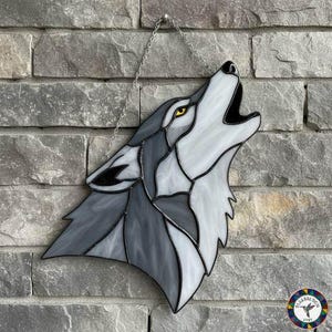 Puede incluir: Lobo aullando de vidriera con secciones grises, blancas y negras, ojos amarillos y una cadena plateada para colgar. El lobo está contra una pared de ladrillos grises.