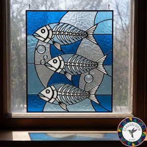 May include: Stained glass panel featuring three stylized fish skeletons in shades of blue, gray, and white. The design includes bubbles and is set against a blue and gray background. The panel is framed in black and hangs in a window.