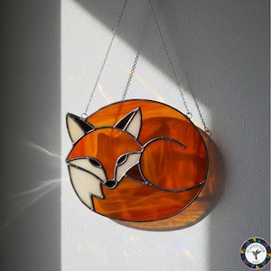 May include: Stained glass fox suncatcher. The fox is curled up in a circle, with orange and white glass. The fox has black accents and is hanging from a silver chain. The suncatcher is a decorative item.