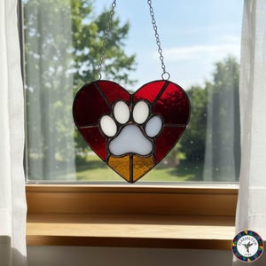 Pode incluir: Coração de vitral com uma pegada branca, detalhes em vermelho, branco e amarelo, pendurado por uma corrente. A decoração em forma de coração é projetada para ser pendurada em uma janela, ideal para amantes de animais.