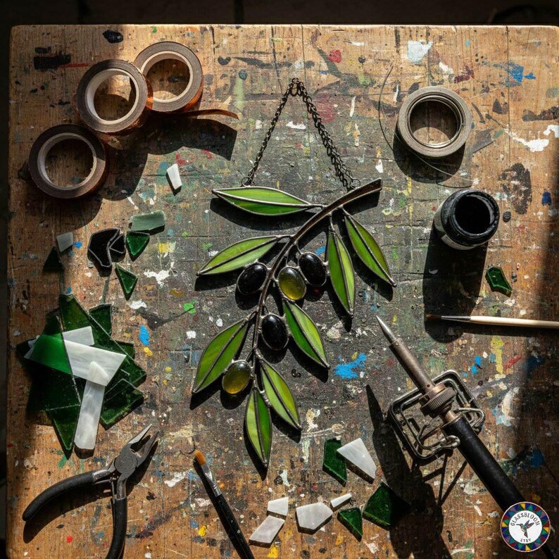 May include: A stained glass olive branch with green leaves and black and green olives hangs from a chain. Various tools and materials, including pliers, solder, and tape, are scattered on a wooden work surface, suggesting a craft project.