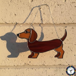 May include: A stained glass dachshund dog suncatcher with a brown and orange color scheme. The dog is suspended by a silver chain. The suncatcher is hanging in front of a light-colored brick wall.