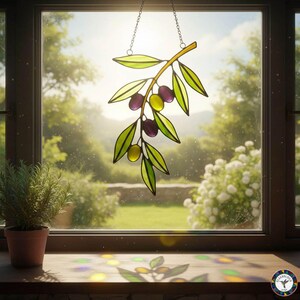 May include: Stained glass olive branch window hanging. The branch is yellow and green with purple and yellow olives. The leaves are outlined in black. The piece hangs from a chain. The window is in front of a sunny outdoor scene.