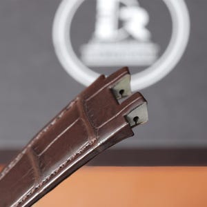 Puede incluir: Primer plano de una correa de reloj de cuero marr&oacute;n con un patr&oacute;n de cocodrilo. La correa tiene forma rectangular con un extremo de hebilla. El fondo est&aacute; borroso, con un logotipo circular blanco.