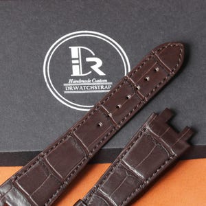 Puede incluir: Primer plano de una correa de reloj de cuero marr&oacute;n con un patr&oacute;n de piel de cocodrilo. La correa tiene un borde cosido y varios agujeros para ajustar. El logotipo de la marca es visible en el fondo.