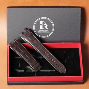 Puede incluir: Un juego de correas de reloj de cuero marr&oacute;n, presentado en una caja negra con interior rojo. Las correas del reloj tienen una textura de piel de cocodrilo. La tapa de la caja presenta un logotipo circular blanco con el texto "Handmade Custom DRWATCHSTRAP".
