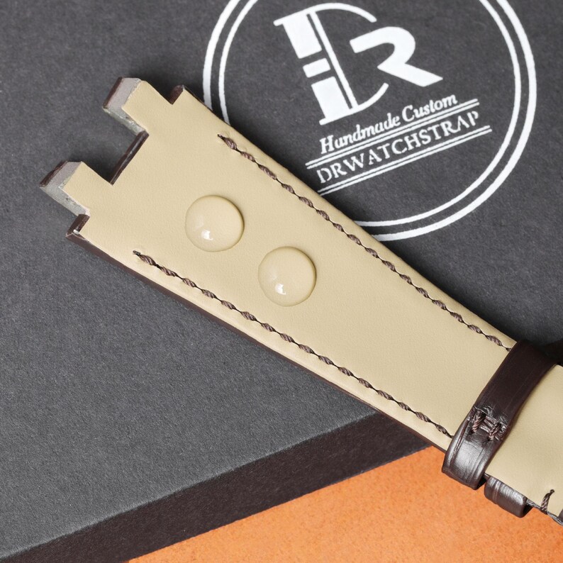 Puede incluir: Primer plano de una correa de reloj de cuero color beige con un acento marr&oacute;n oscuro. La correa presenta dos tachuelas redondas de color claro y est&aacute; cosida con hilo oscuro. El texto "Handmade Custom DRWATCHSTRAP" es visible en el fondo.