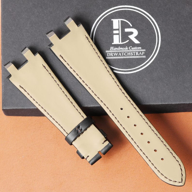 Op de afbeelding: Tweedelig horlogebandje van beige leer met zwarte stiksels. De band heeft een zwarte leren lus en een roestvrijstalen gesp. De tekst "Handmade Custom DRWATCHSTRAP" is zichtbaar op de verpakking.