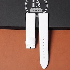 May include: Two white leather watch straps with a textured finish. One strap features a buckle, and the other has adjustment holes. The straps are set against a brown and black backdrop. The logo "Handmade Custom DRWATCHSTRAP" is visible.