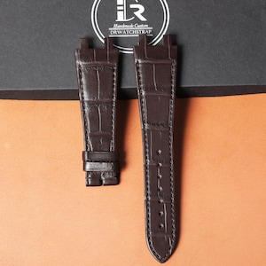 May include: Two dark brown leather watch straps with a crocodile skin texture. One strap has a buckle, and the other has several holes for adjustment. The straps are displayed on a brown and black background.