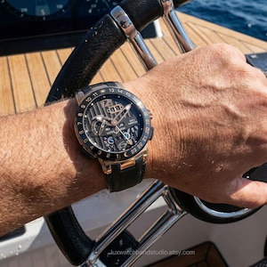 Op de afbeelding: Een luxe polshorloge met een zwarte en gouden wijzerplaat en een zwarte leren band. Het horloge heeft meerdere wijzerplaten en een datumweergave. Het horloge zit om een pols, met een houten dek en water op de achtergrond.