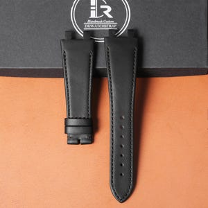 May include: Two black leather watch straps with stitching detail. One strap has a buckle, the other has multiple holes. The straps are displayed on a brown surface with a black box in the background. The text "Handmade Custom DRWATCHSTRAP" is visible.