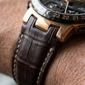 Puede incluir: Primer plano de un reloj de lujo con correa de cuero marr&oacute;n. La esfera del reloj es negra con detalles dorados y una esfera detallada. La correa presenta un patr&oacute;n texturizado, similar al de un cocodrilo, con costuras blancas.