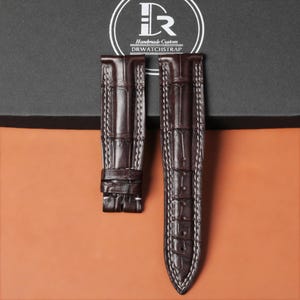 Puede incluir: Dos correas de reloj de cuero marrón oscuro con costuras blancas. Las correas tienen una textura de piel de cocodrilo. La marca "DRWATCHSTRAP" está impresa en la parte superior de la imagen.