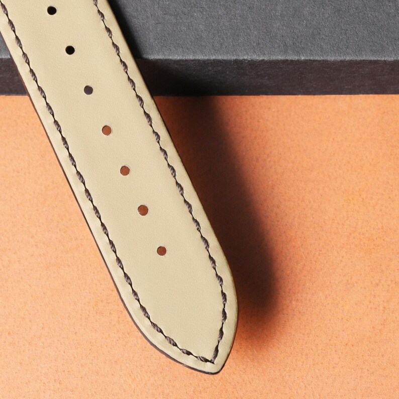 Puede incluir: Primer plano de una correa de reloj de cuero color crema con costuras marr&oacute;n oscuro y agujeros espaciados uniformemente. La correa est&aacute; en &aacute;ngulo, mostrando el detalle de las costuras y la textura del cuero. El fondo es naranja y gris.