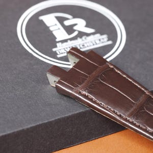 Puede incluir: Primer plano de una correa de reloj de cuero marr&oacute;n con textura de piel de cocodrilo. La correa es parcialmente visible, con los extremos de los cierres met&aacute;licos a la vista. El fondo presenta una caja negra con un logotipo y texto blancos.