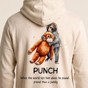 Op de afbeelding: Beige hoodie met een aquarel illustratie van twee omhelzende apen. Eén is oranje, de ander grijs. Het woord "PUNCH" staat onder de afbeelding, samen met de tekst: "Toen de wereld hem alleen liet, vond hij een vriend in plaats van een familie."
