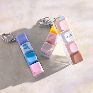 May include: Two keychains featuring colourful, square charms. One has a purple, white, blue, and pink charm with a magnifying glass icon. The other has pink, white, and brown charms, one with a smiling face. Both have silver key rings.