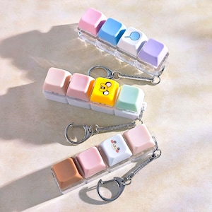 May include: Three keychains featuring colourful keyboard keycaps. Each keychain has a different colour scheme, including pink, blue, yellow, and white. Some keycaps feature small illustrations. The keychains have silver-coloured hardware.