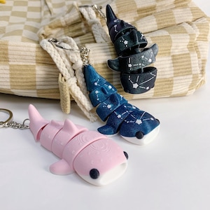 Puede incluir: Tres llaveros articulados de tiburón ballena en rosa, azul y negro. El tiburón rosa tiene una barriga blanca y ojos negros. Los tiburones azules y negros presentan un diseño de constelación. Todos los llaveros tienen llaveros plateados.