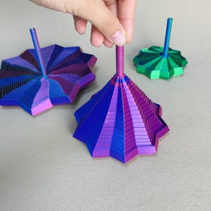 May include: Three colorful, metallic spinning tops. The largest is held by a hand and features a gradient of blue, purple, and pink. Two others are visible, one in similar colors and another in green and blue.