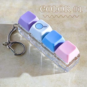 May include: A keycap keychain with pastel-coloured keycaps in a clear plastic holder. The keycaps are purple, white with a pan graphic, blue, and pink. The text "COLOR #1" is above the keycaps.