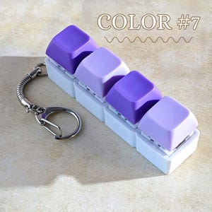 May include: A keychain with five purple keyboard keycaps in different shades, mounted on a white base. It has a silver metal chain and clasp. The text "COLOR #7" is above the keycaps. The keycaps are in shades of purple.
