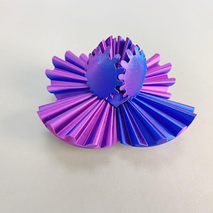 Op de afbeelding: Een 3D-geprinte, abstracte sculptuur in tinten paars en blauw. Het ontwerp heeft een centrale, tandwielachtige structuur, omgeven door gelaagde, bloembladachtige elementen. De kleurovergang verschuift van paars naar blauw, waardoor een opvallend stuk ontstaat.