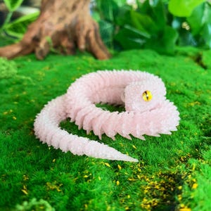 May include: A pink, segmented, worm-like sculpture with a textured surface and a yellow eye. The sculpture is coiled on a bed of green moss, with a blurred background featuring a tree trunk and foliage. The worm's body has a gradient from pink to white.