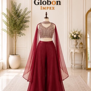 May include: A maroon two-piece outfit on a mannequin featuring a sleeveless crop top with intricate embroidery and a matching choker collar. The ensemble includes wide-leg trousers and a sheer, floor-length cape attached at the shoulders.