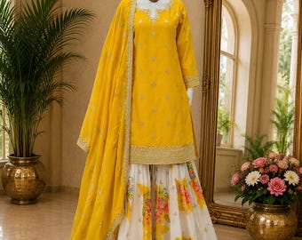 May include: A bright yellow long-sleeved tunic with silver embroidery paired with a white floor-length skirt featuring a colorful floral print and a wide gold embroidered hem. A matching yellow dupatta with silver accents is draped over the shoulder.