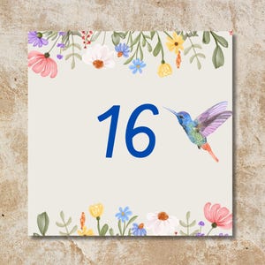Può includere: Una scheda quadrata con il numero 16 in blu, un colibrì ad acquerello e decorazioni floreali. La scheda ha uno sfondo beige chiaro e un bordo di fiori. La parte inferiore della scheda ha il testo: "Choisissez votre numéro et nom ou mot personnalisé."